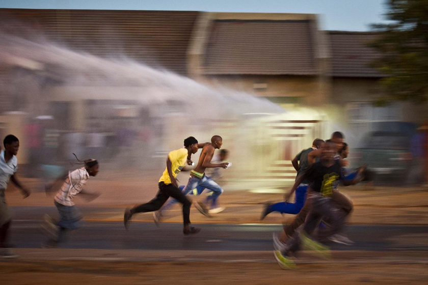 2012年3月19日，南非Ratanda，示威者逃离警察的水枪喷射。抗议电力中断的示威者点燃了市政局办公室旁边的两间房屋，并与警察发生冲突。 Gallo Images/Getty Images/CFP_财新每周图片(2012.3.17-3.23) 