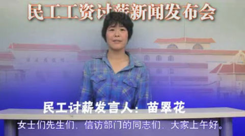 （视频截图）这名“民工讨薪发言人”苗翠花，身着蓝色方格衬衣的中年妇女在镜头前，在她身后的背景板上写着“民工工资讨薪新闻发布会”的字样。